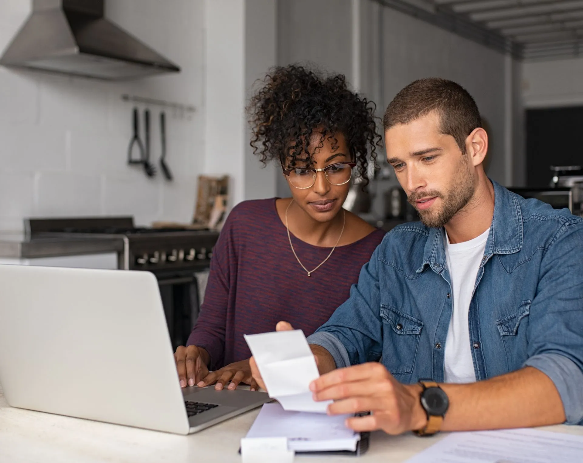 A couple sits together at a dining table, both focused on bills and papers in front of them. The couple is in deep discussion, one of them typing on the laptop while the other reviews the paperwork.