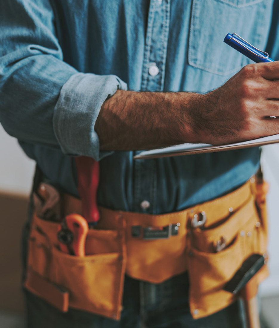 Close-up of a male contractor hand, writing on a clipboard. His leather toolbelt can be seen with various tools in it.