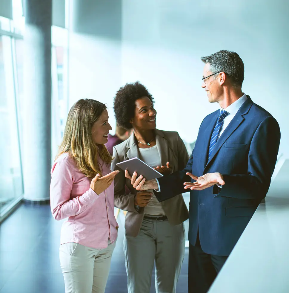 Businessman in suit holding tablet talking with two women standing in building lobby.